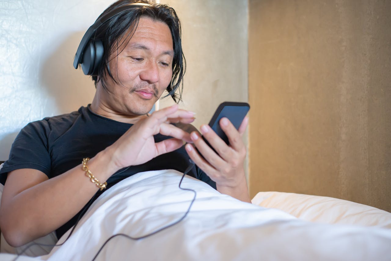 Asian man enjoying leisure time in bedroom using smartphone and headphones.